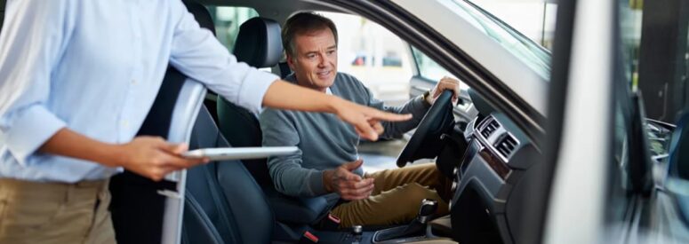 A man sitting in a car being instructed on how to use the vehicle by a woman with a tablet